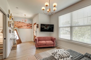 A spacious living room with a large window, a pink sofa, a mounted TV, and a painting on the wall. - 4 Mi to Broadway: Chic Townhome w/ Home Office! (Nashville)
