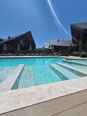 A swimming pool with clear water, surrounded by lounge chairs and umbrellas, with buildings in the background.