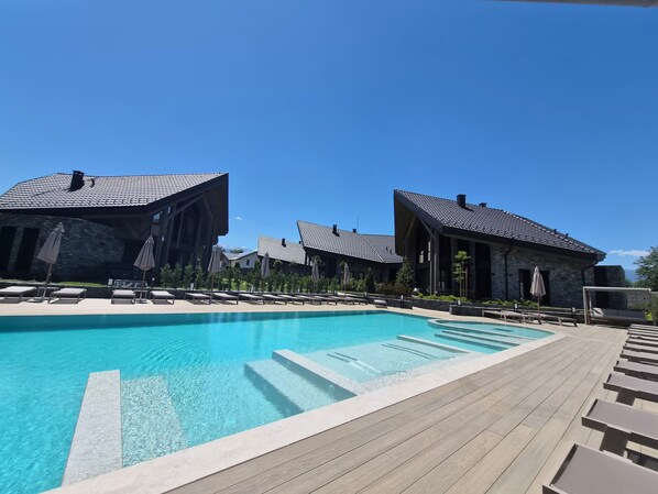A poolside area with lounge chairs and umbrellas, surrounded by stone buildings with sloped roofs.