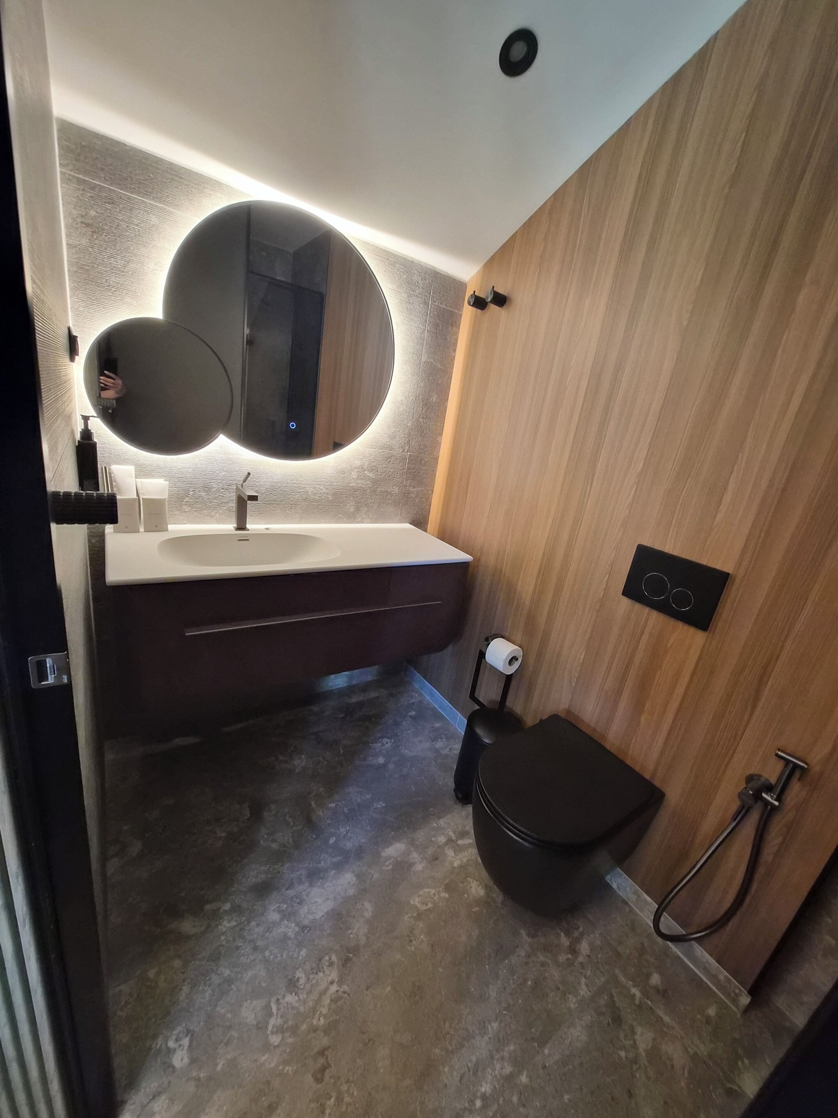 Une salle de bain moderne avec un miroir rond, un lavabo blanc, des toilettes de couleur sombre et un espace douche.