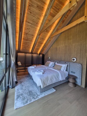 A bedroom with a sloped ceiling, two beds, a bedside table, and a large window with curtains.