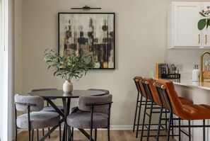 Uma sala de jantar com uma mesa redonda, quatro cadeiras, um vaso com uma planta e um quadro na parede.
