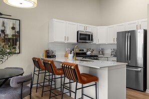 A modern kitchen with white cabinets, stainless steel appliances, and a central island with bar stools. - Luxury 2BR/2BA Condo |Walk to ND| Private Balcony (South Bend)