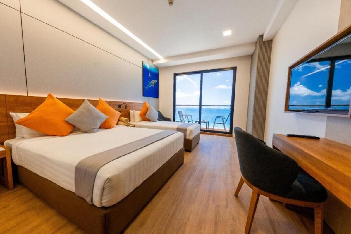 Habitación de hotel con una cama grande, un escritorio, una silla y vistas al océano a través de un ventanal amplio.