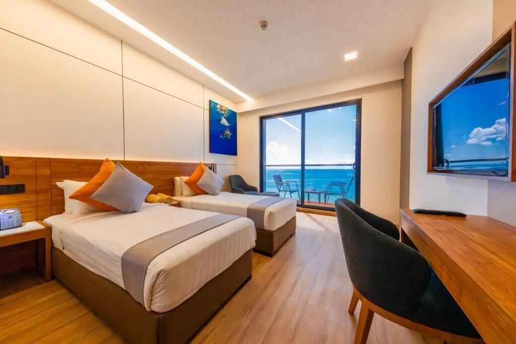Habitación de hotel con dos camas, un escritorio, una silla, un televisor y un amplio ventanal con vista al mar.
