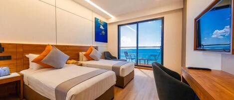 A hotel room with two beds, a desk, a chair, a TV, and a large window with a view of the sea.