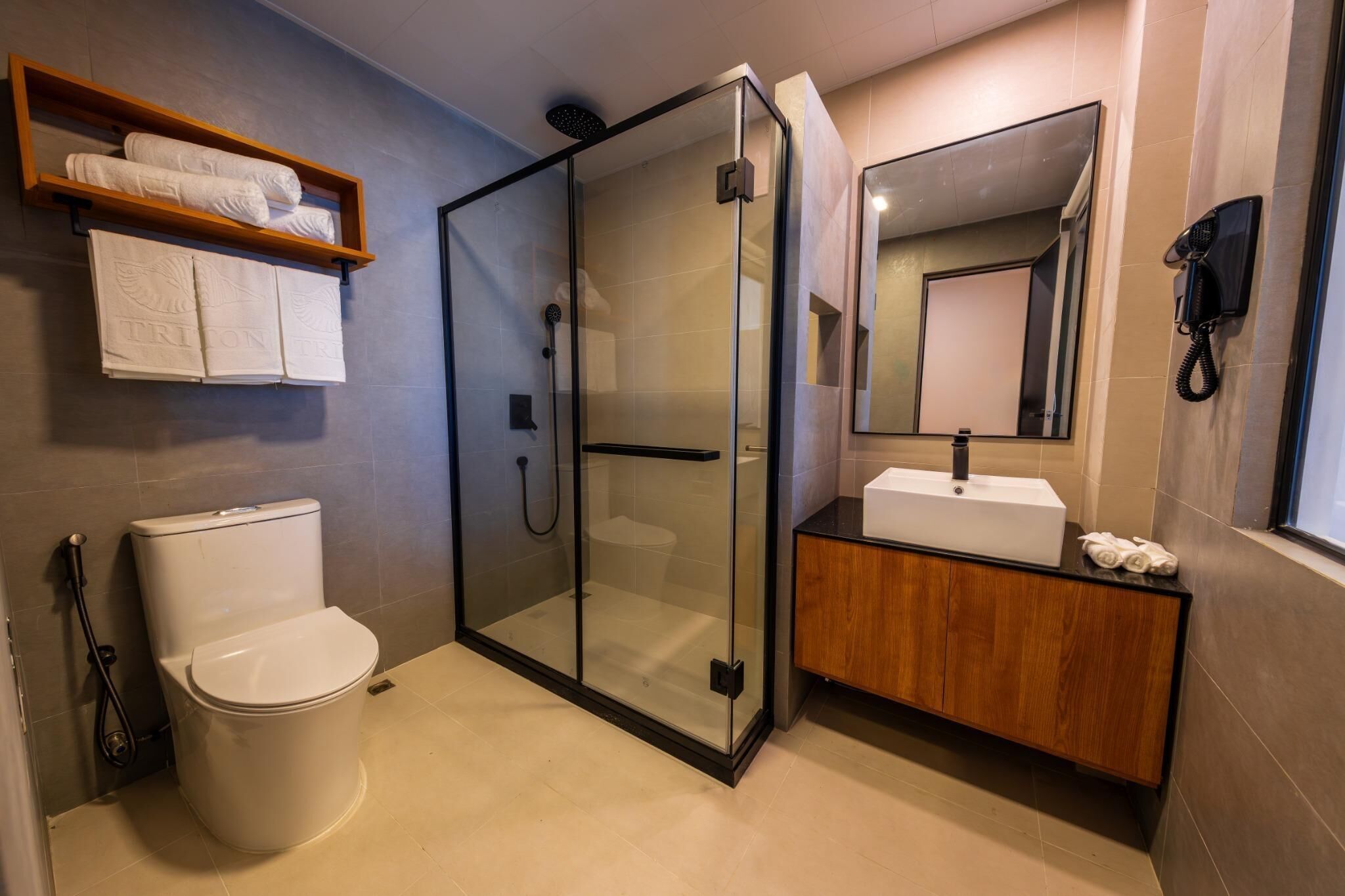 Un baño moderno con ducha de vidrio, inodoro blanco, tocador de madera con lavabo y un perchero sobre el inodoro.