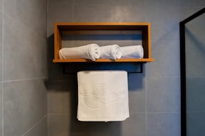 A wooden shelf with two rolled towels and a towel with a sea shell design on a tiled wall. - Triton Exotica (Maafushi)