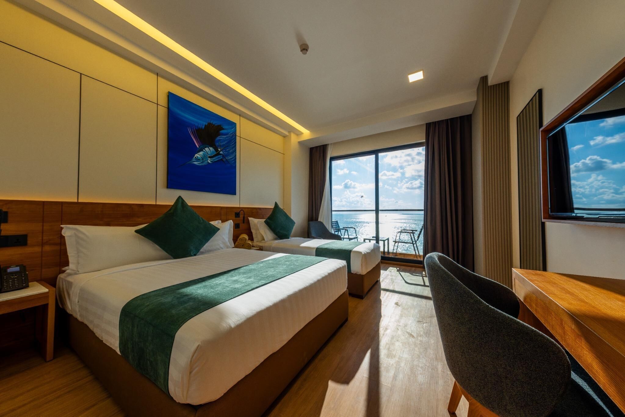 A hotel room with two beds, a desk, a chair, a large window with a view of the sea, and a painting on the wall.