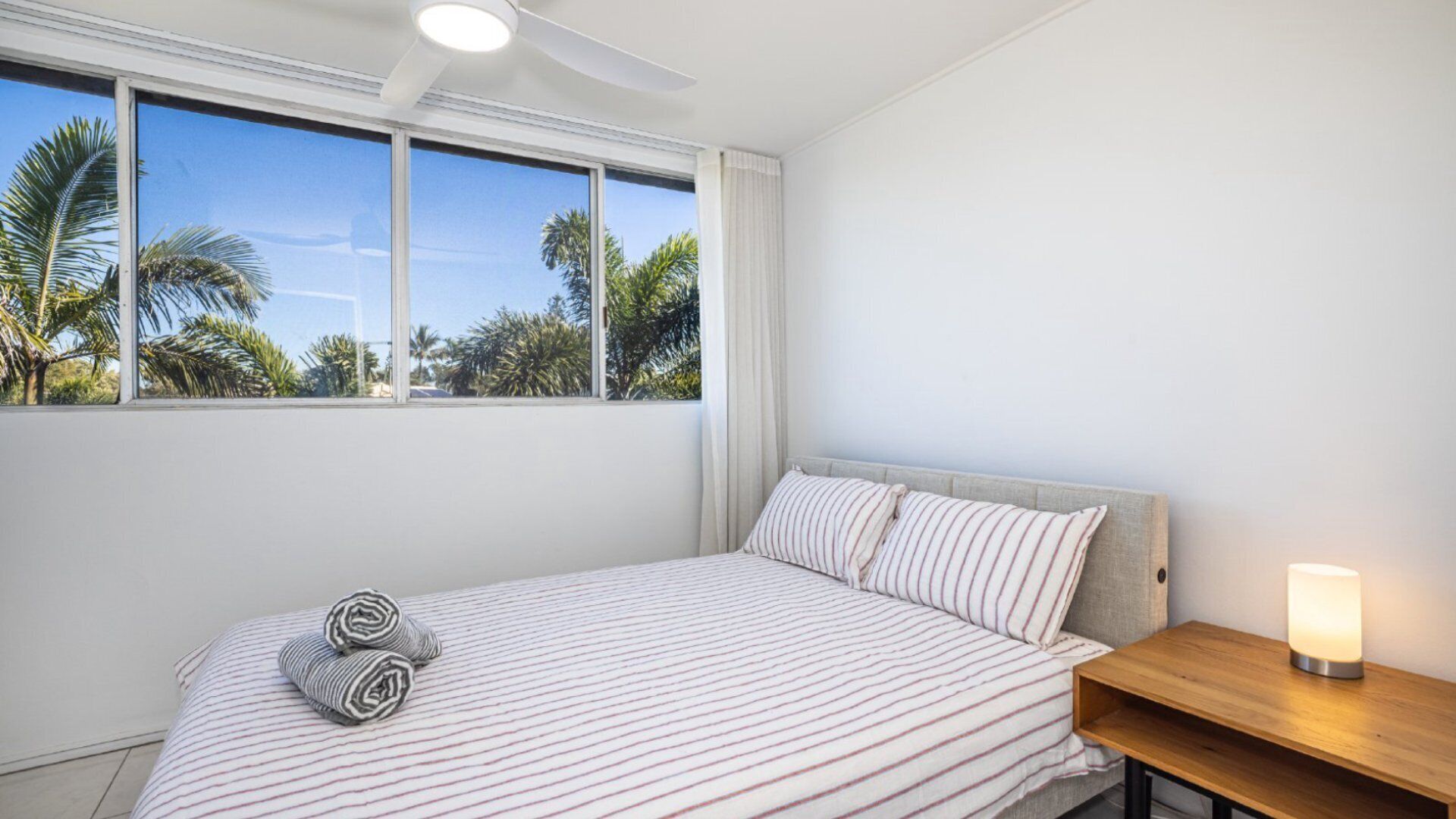 A bedroom with a bed, a nightstand, a lamp, and a window with a view of palm trees.