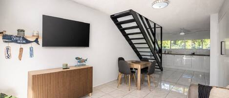 A modern living room with a flat-screen TV, a wooden console, a dining area with chairs, and a kitchen in the background.