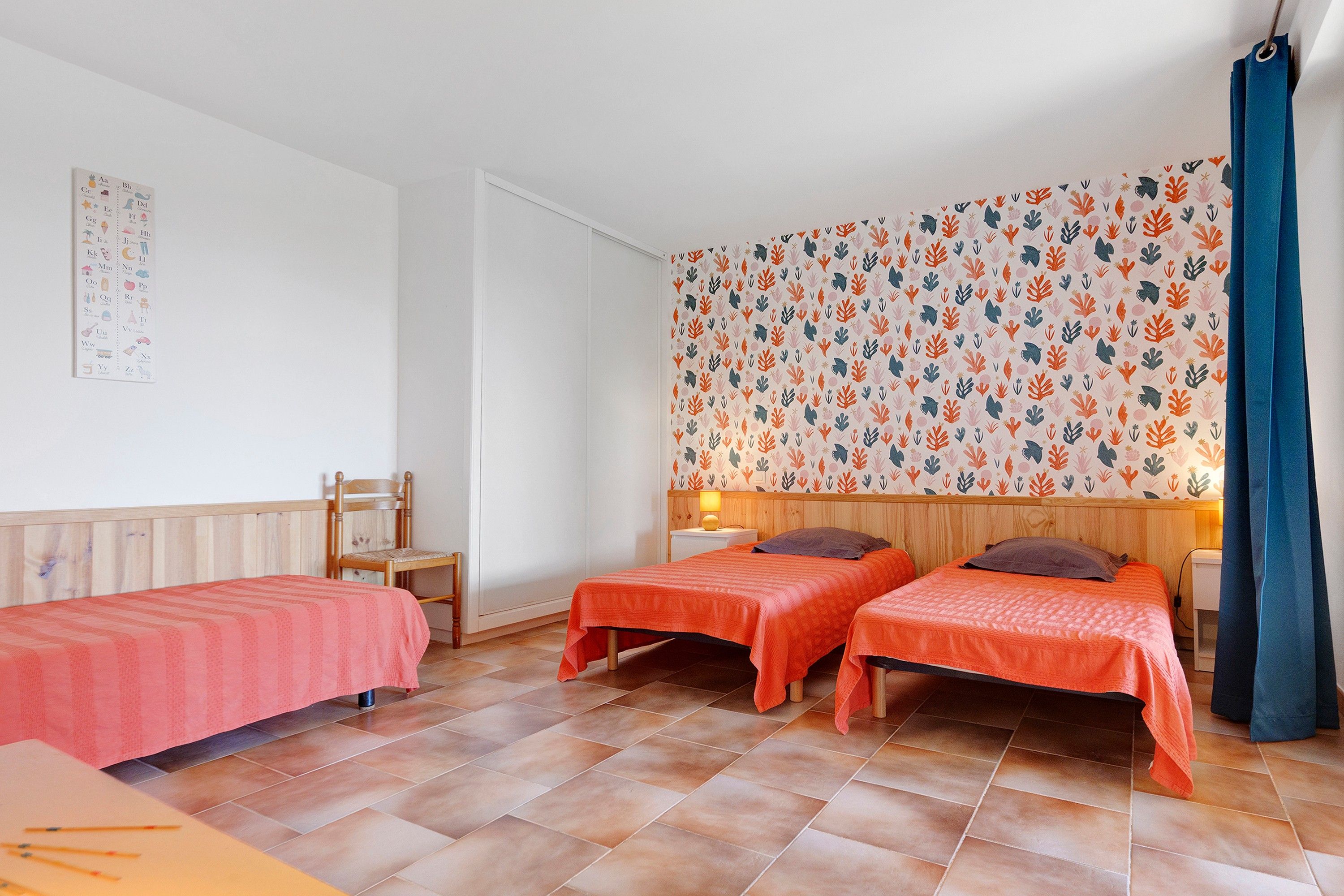 A room with two single beds, a wooden headboard, a small table, and a wall with a colorful plant pattern.