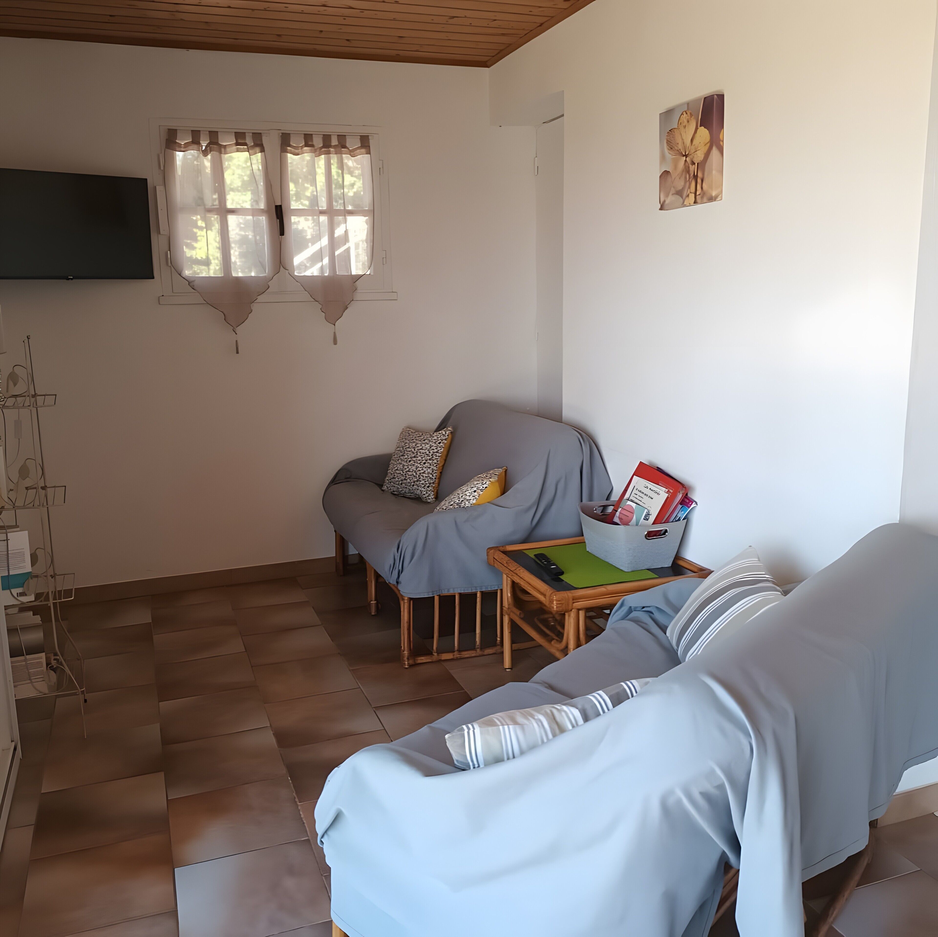 Ein gemütliches Zimmer mit einer Couch, einem kleinen Tisch mit einem Korb, einem an der Wand befestigten Fernseher und einem Fenster mit Vorhängen.