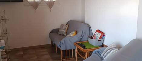 Ein gemütliches Zimmer mit einer Couch, einem kleinen Tisch mit einem Korb, einem an der Wand befestigten Fernseher und einem Fenster mit Vorhängen.