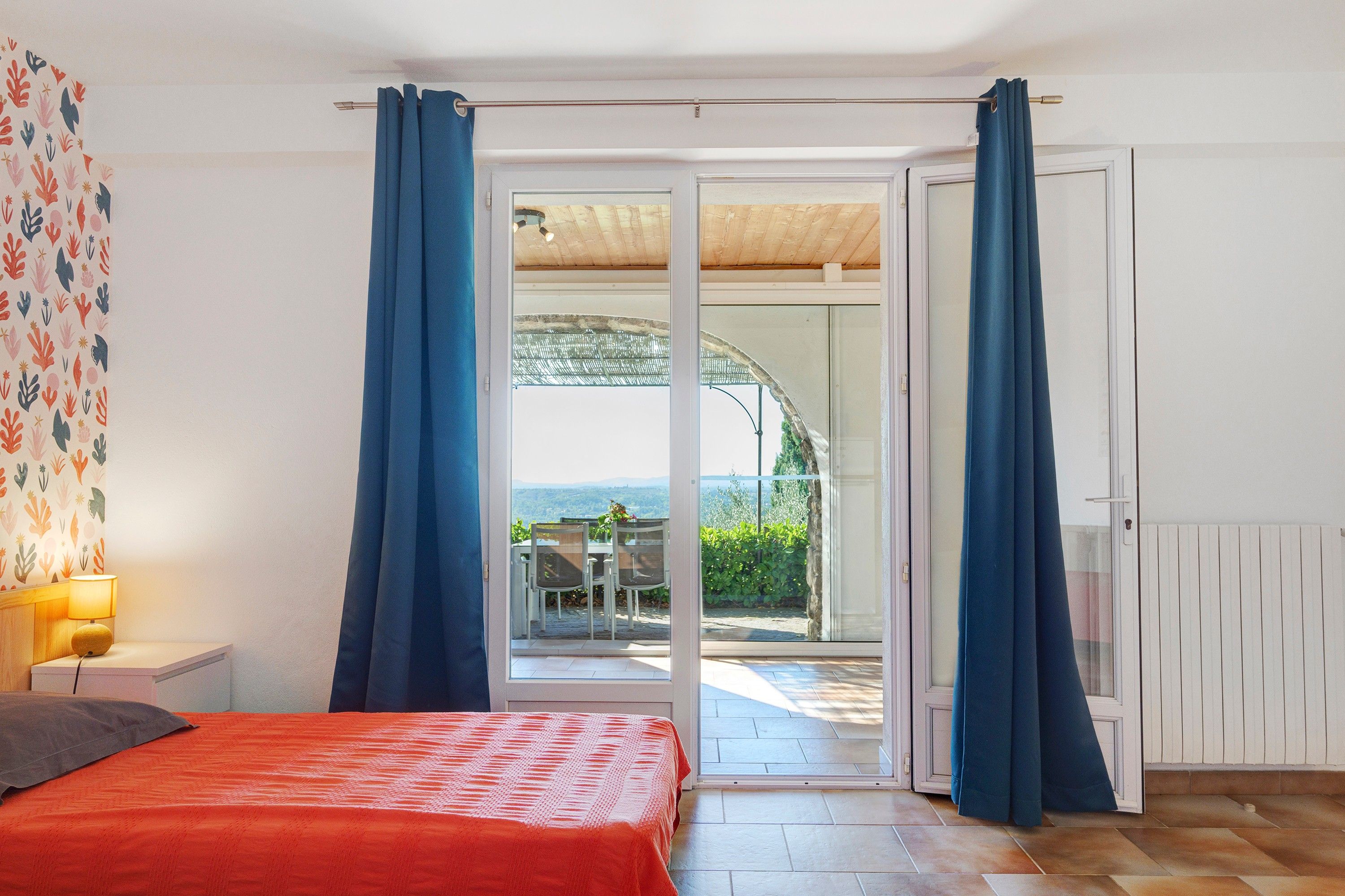 Ein Schlafzimmer mit Bett, Nachttisch, Lampe und Blick auf eine Terrasse mit Tisch und Stühlen.