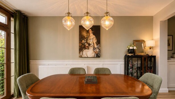 Une salle à manger avec une grande table en bois, quatre chaises rembourrées, un lustre et un tableau accroché au mur.
