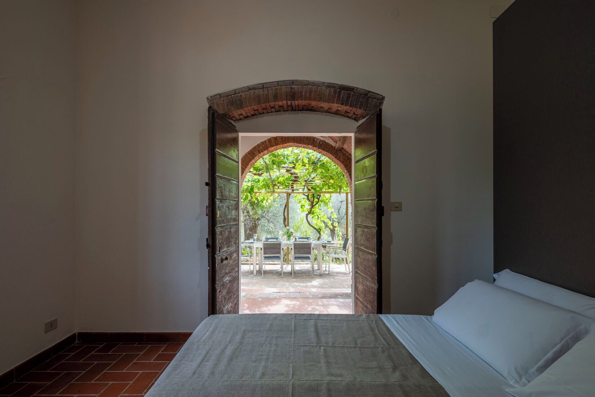 Une pièce avec un lit, un arc en briques donnant sur un espace extérieur aménagé avec une table et des chaises.