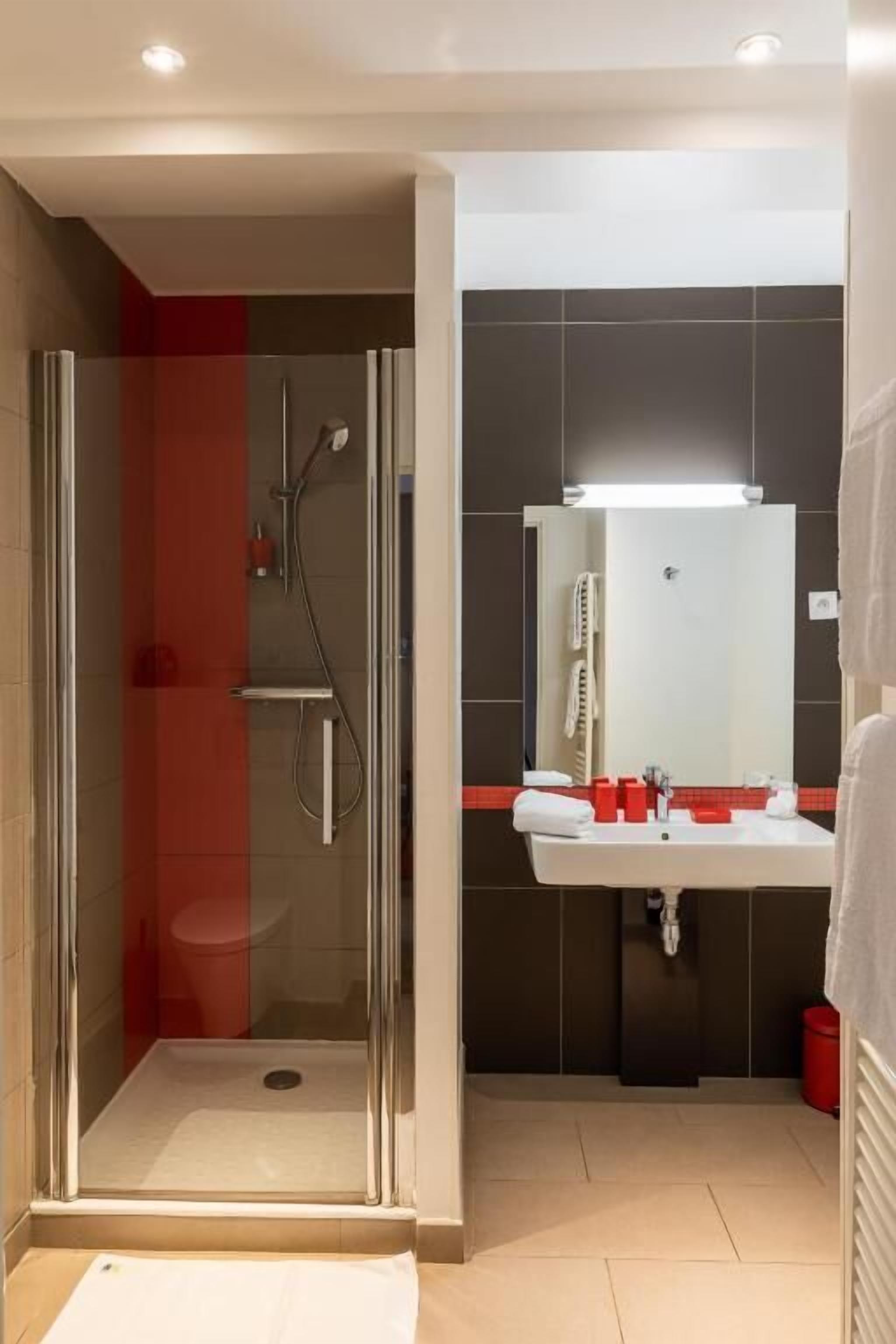 A modern bathroom with a glass shower enclosure, a white sink with red accents, and a large mirror above the sink.