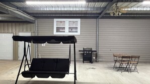 Ein überdachter Sitzbereich mit einer Hängesitzbank, einem Grill und einem Tisch mit Stühlen in einer Lagerhaus-ähnlichen Umgebung.