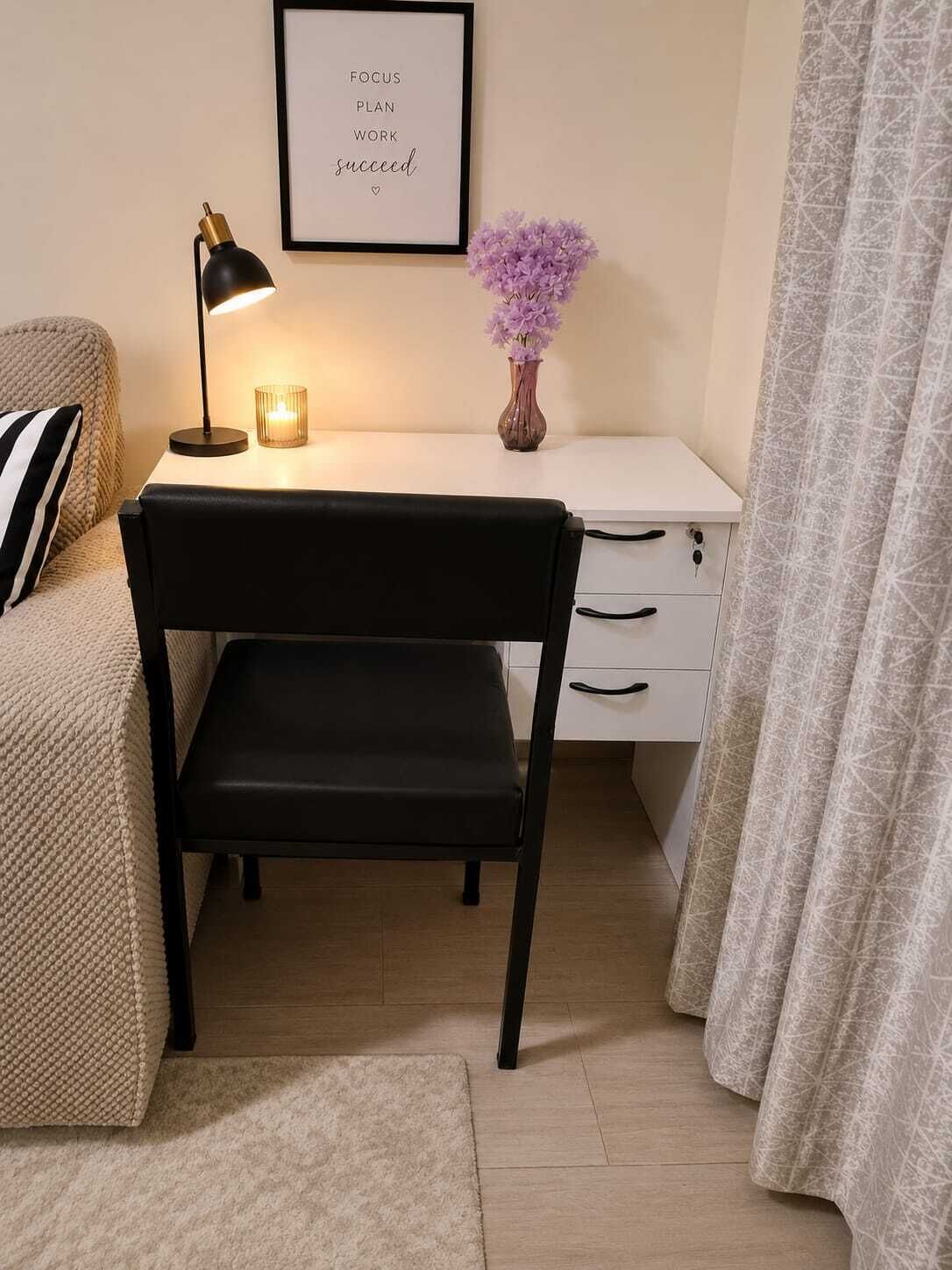Un dormitorio con cama, un escritorio y una silla, una lámpara de noche, una cita motivacional enmarcada y un florero con flores.