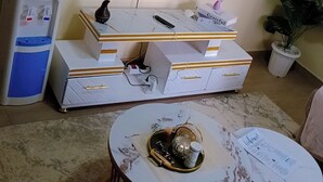 Een woonkamer met een flatscreen televisie aan de muur, een waterdispenser, een witte kast met lades, een ronde salontafel met dienblad en een potplant.