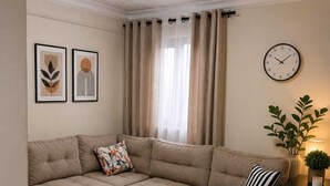 Een woonkamer met een hoekbank, twee ronde bijzettafels, een wandklok en een raam met gordijnen.