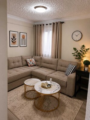 A living room with a sectional sofa, two round coffee tables, a wall clock, and a window with curtains. - Cozy,1-bedroom apartment in a fabulous and quiet Ruiru neighborhood (Ruiru)