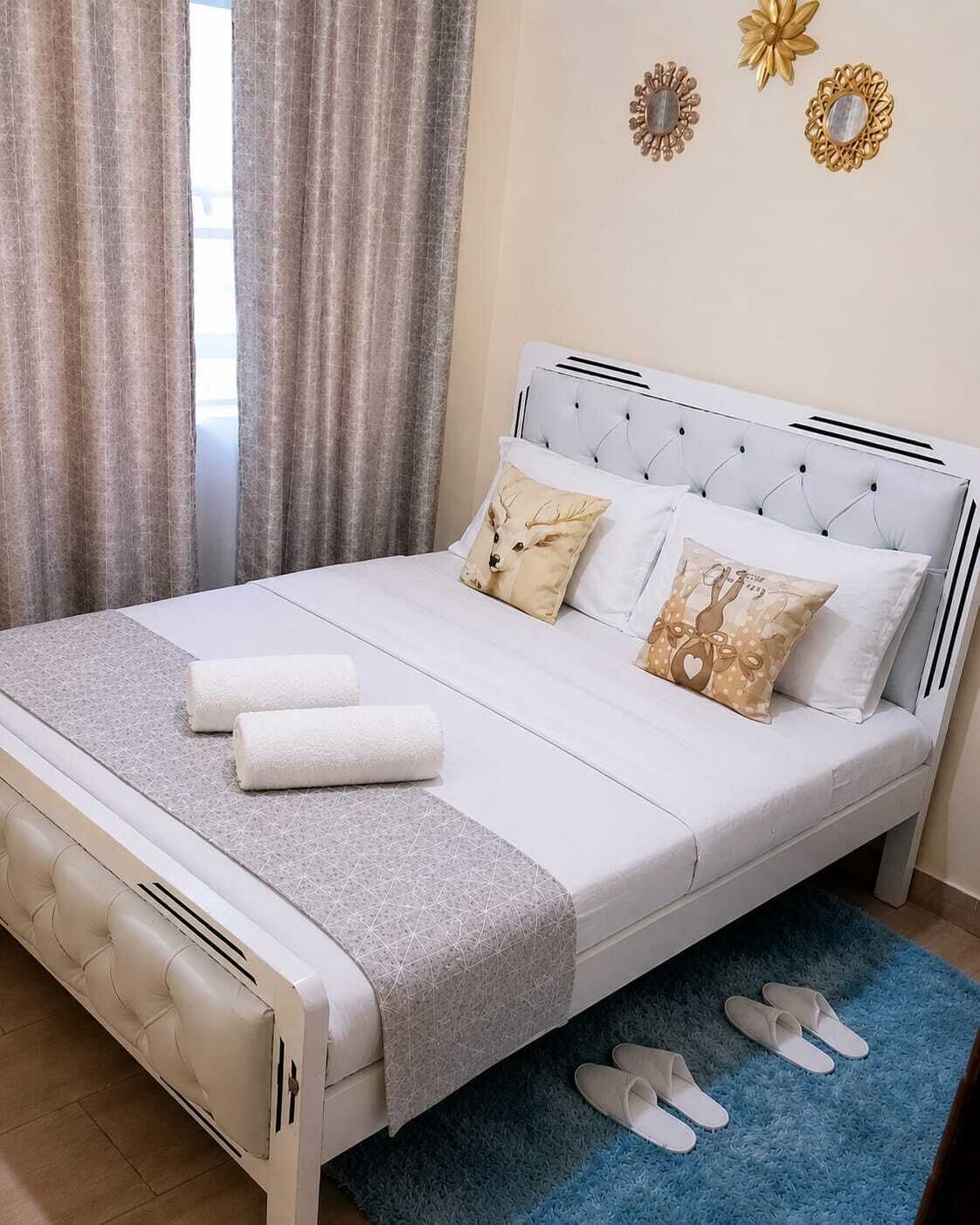 Una cama con cabecera blanca, dos almohadas, una colcha gris y dos toallas enrolladas.