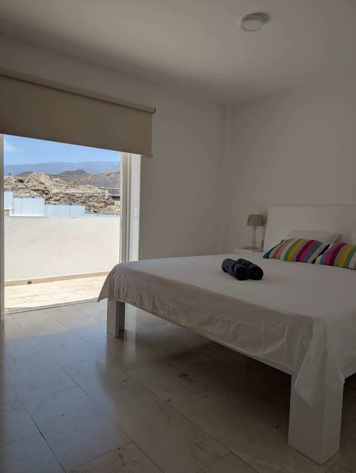 A bedroom with a bed, a lamp, a colorful pillow, and a view of a rocky landscape through a large window.