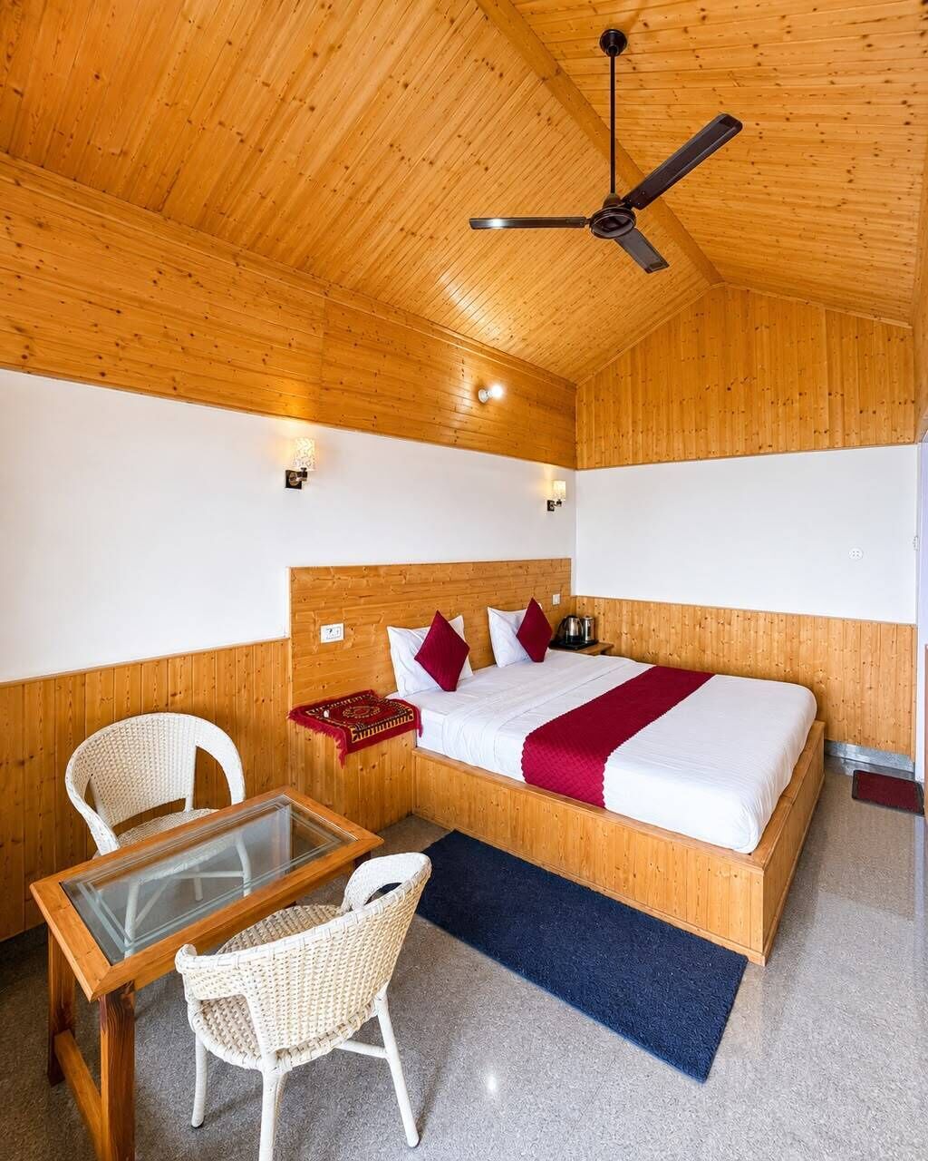Una habitación con paneles de madera, una cama, dos sillas, una mesa y un ventilador de techo.