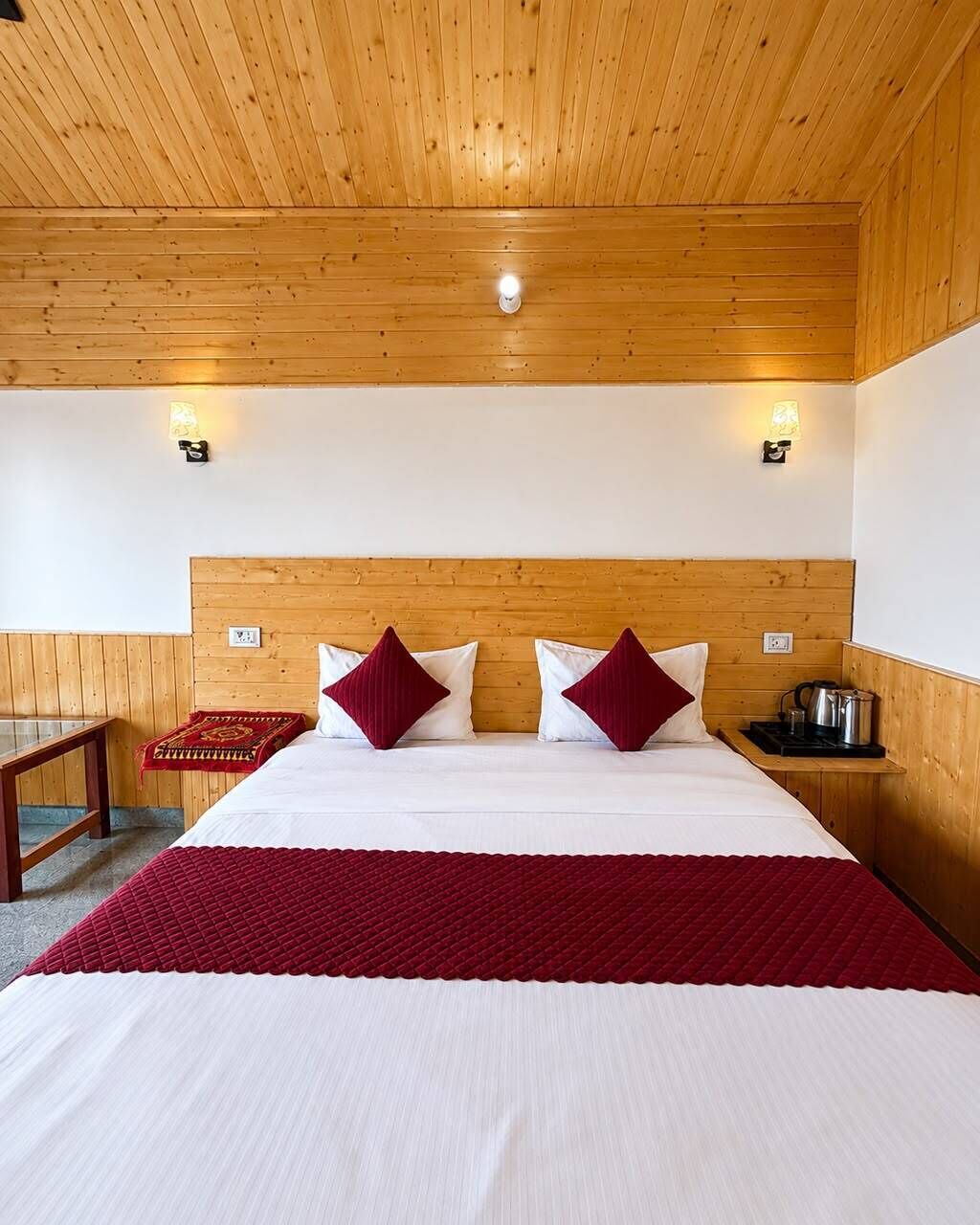 A hotel room with a wooden ceiling, a bed with white and red bedding, two wall-mounted lights, and a small table with a kettle.