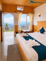 A hotel room with a bed, a wooden headboard, a nightstand, a window with curtains, a view of mountains, and a balcony with a fence.