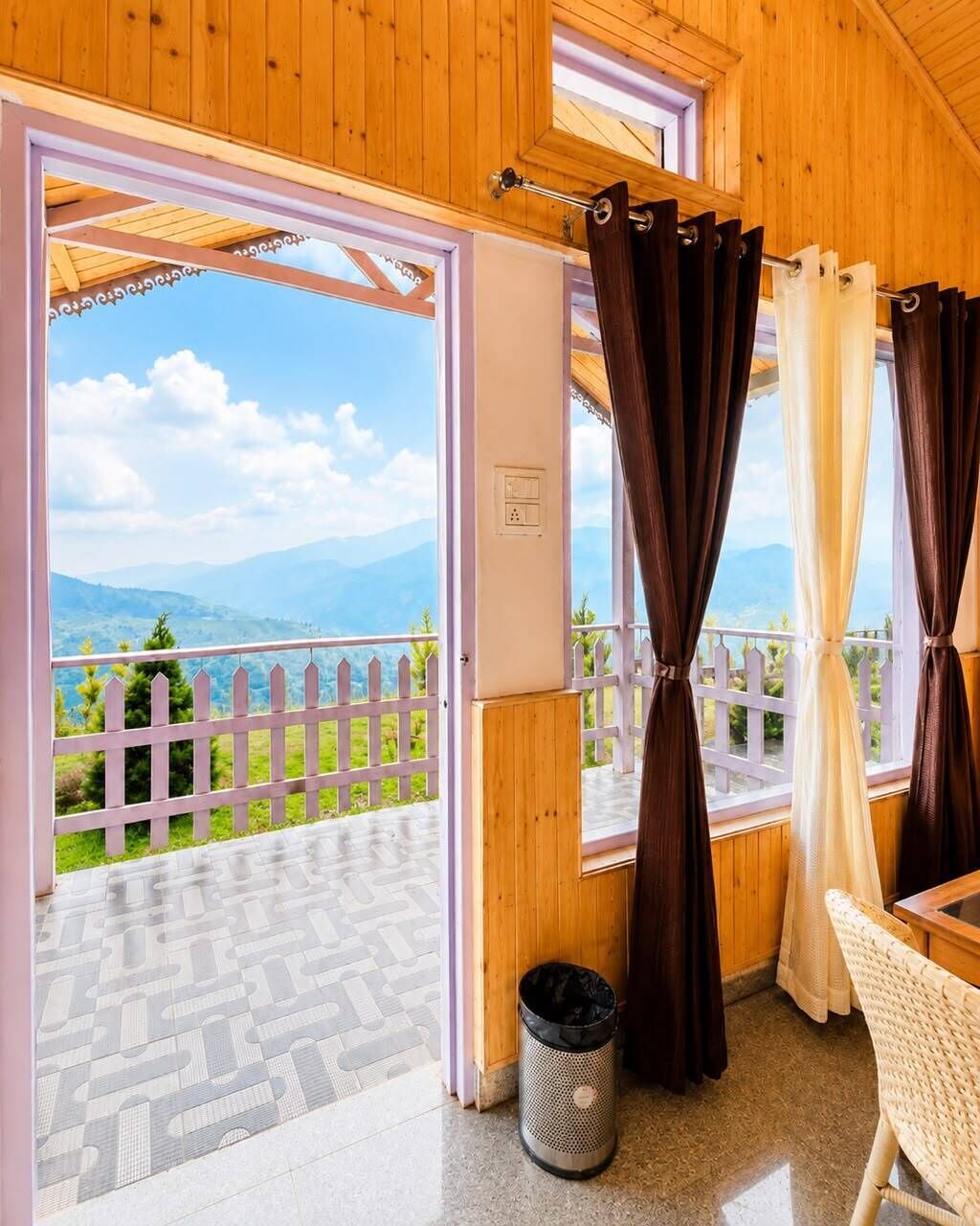 A wooden room with a balcony, a white fence, and a view of green hills.