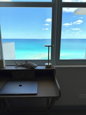 A room with a desk, a laptop, a small table with a lamp, and a view of the ocean through a window. - Oceanfront Gem • Newly Renovated (Miami Beach)