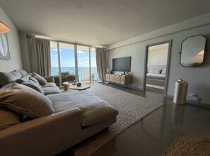 A modern living room with a large sofa, a flat-screen TV, and a view of the ocean through large windows. - Oceanfront Gem • Newly Renovated (Miami Beach)