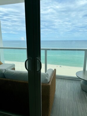 A balcony with a view of the ocean and beach, wicker furniture, and a glass door. - Oceanfront Gem • Newly Renovated (Miami Beach)
