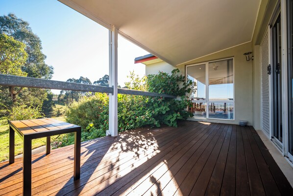 A wooden deck with a table, a covered area with a railing, and a view of greenery and a building in the background.