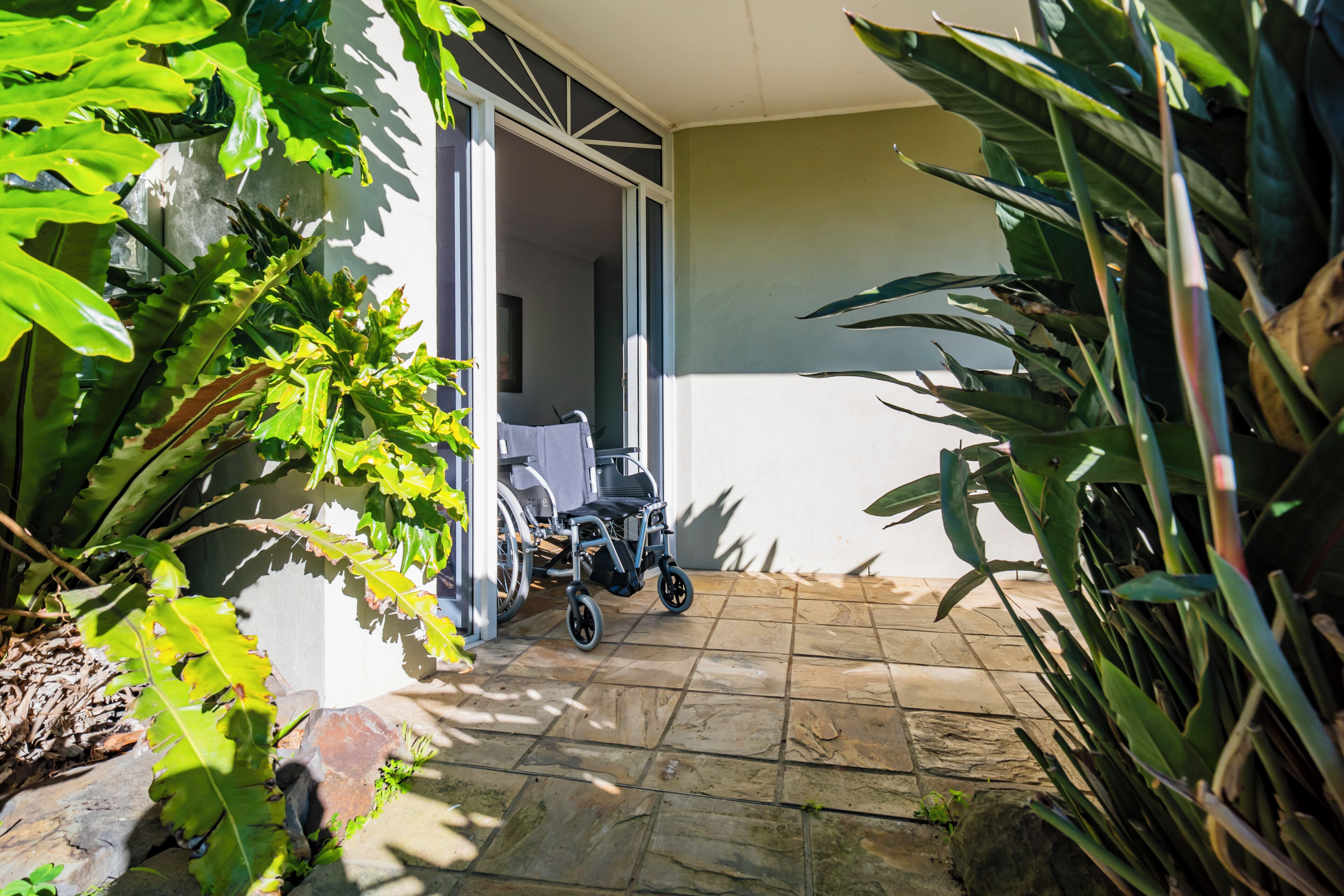 A wheelchair in a covered outdoor area with a tiled floor and large green plants.
