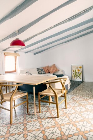 A room with a wooden dining table, chairs, a sofa with patterned cushions, a red pendant light, and a framed botanical print on the wall. - A charming little house nestled between vineyards and the sea. (Saifores)