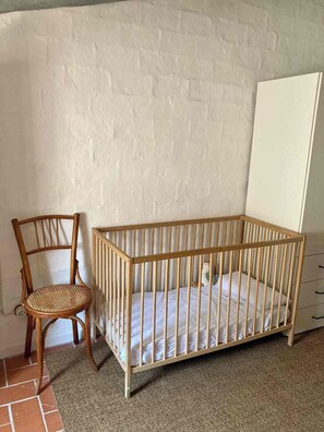 A wooden crib with a mattress, a wicker chair, and a white wardrobe in a room with a white brick wall. - A charming little house nestled between vineyards and the sea. (Saifores)