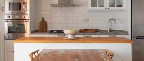 Una cocina con armarios blancos, una isla de madera, una mesa de comedor con sillas y una lámpara colgante de mimbre.