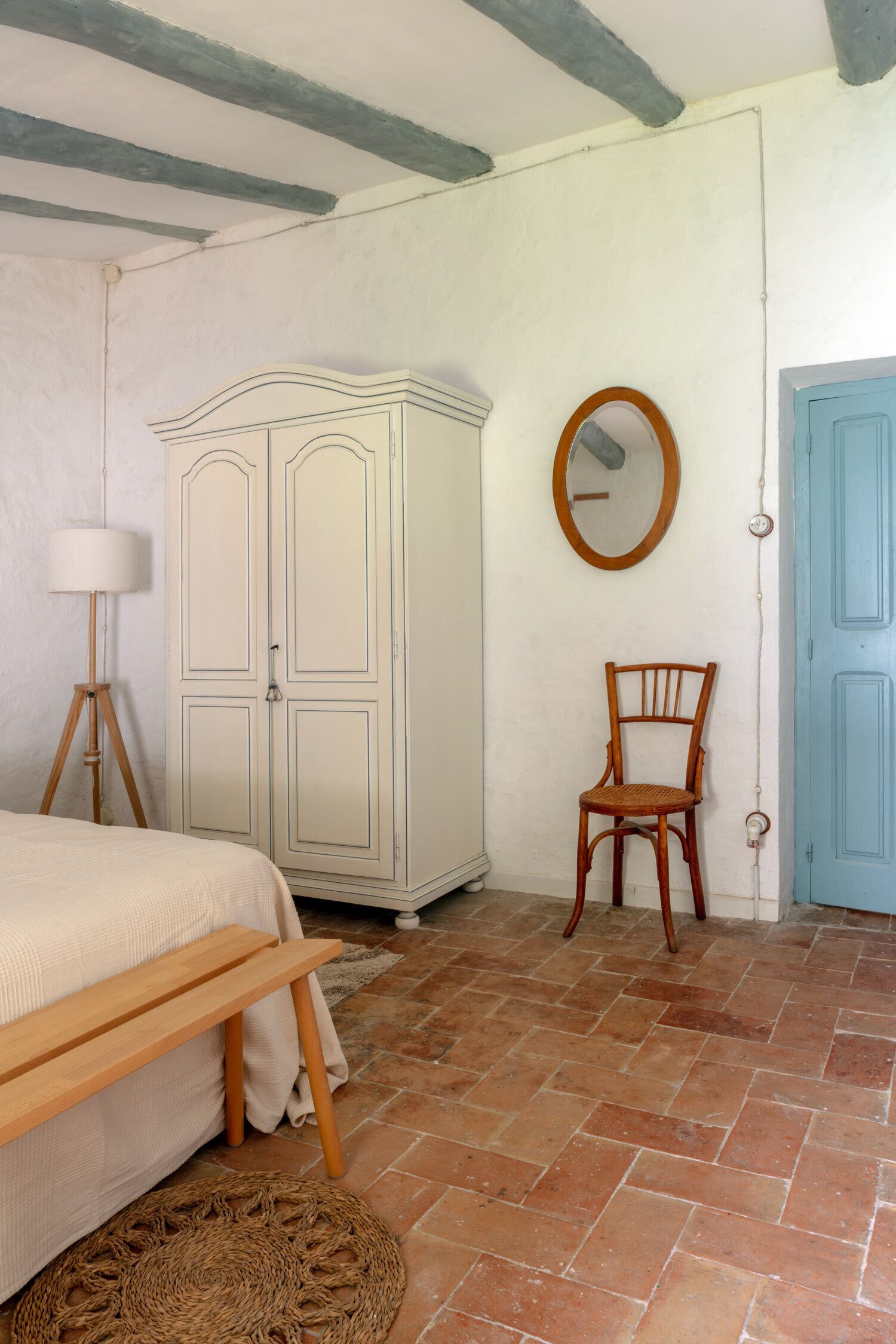 Een slaapkamer met een bed, een houten bankje, een rieten mat, een kledingkast, een stoel, een spiegel en een blauwe deur.