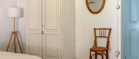 A bedroom with a bed, wooden bench, wicker rug, wardrobe, chair, mirror, and a blue door.