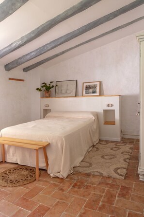 A bedroom with a bed, wooden bench, potted plant, framed pictures, and a small shelf. - A charming little house nestled between vineyards and the sea. (Saifores)