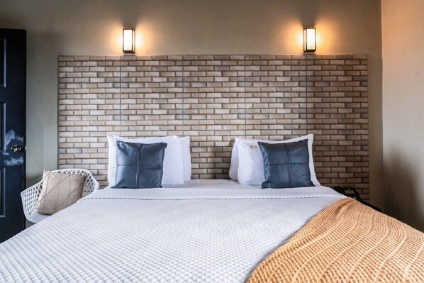 Habitación de hotel con cabecera de ladrillo, dos camas blancas con almohadas grises y blancas, colcha texturizada blanca y una manta tejida.