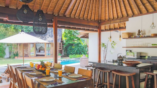 An outdoor dining area with a pool view, wooden furniture, and a kitchen area with stools. - Villa Charlie Seminyak (Bali)
