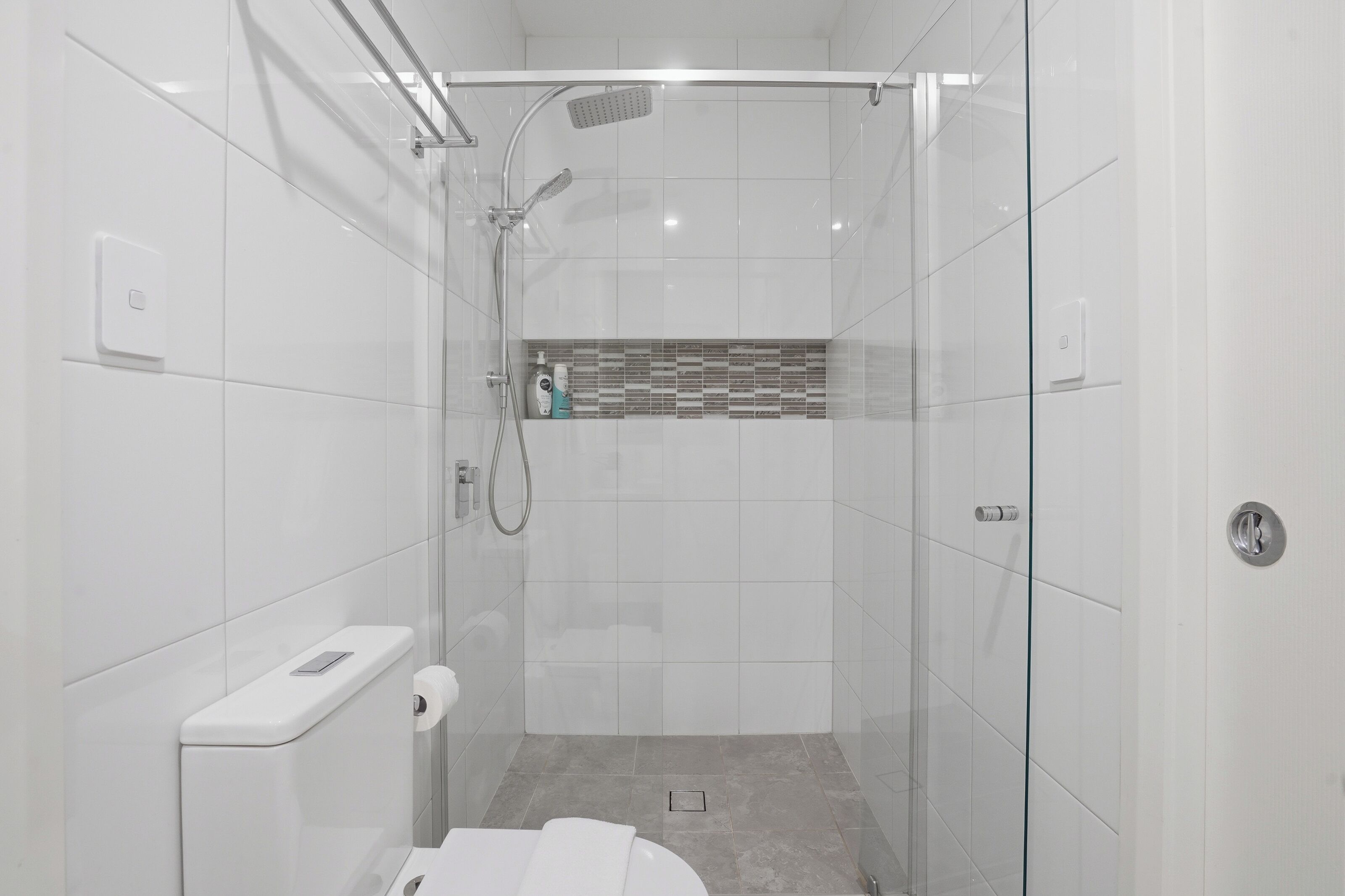 Ein modernes Badezimmer mit einer Duschkabine, weißen Fliesen, einer Toilette und einem kleinen Ablagefach für Pflegeprodukte.
