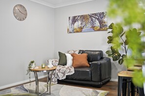 Ein Wohnzimmer mit einem schwarzen Ledersofa, einem Beistelltisch mit Pflanze, einer Uhr und einem Bild von einem Baumweg.