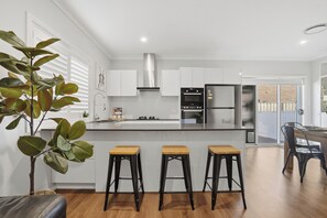 A modern kitchen with white cabinets, a stainless steel range hood, and a central island with bar stools. - The Gledswood Retreat | Stylish 3BR Stay with Space & Privacy (Gledswood Hills)
