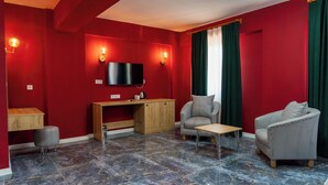Una habitación con paredes rojas, televisor de pantalla plana, un escritorio de madera, dos sillones a cuadros, un otoman redondito y una mesa de centro.